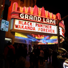 A glowing theater marquee sign reads, "Black Panther: Wakanda Forever". A crowd walks by under the sign. 